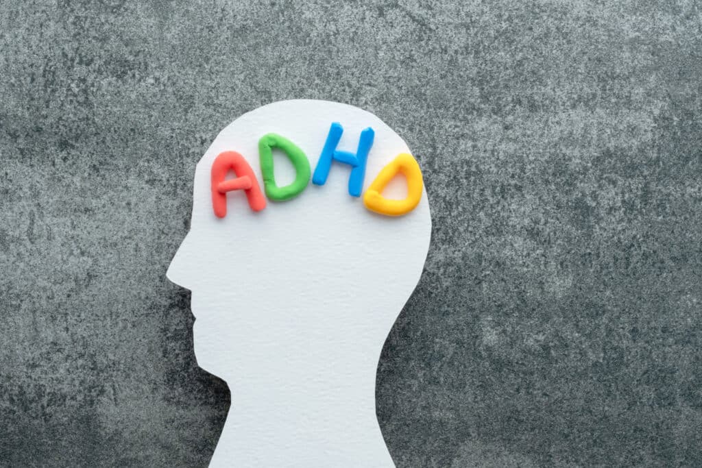 A white silhouette of a human head with colorful letters spelling "ADHD" placed on the brain area, set against a textured gray background.