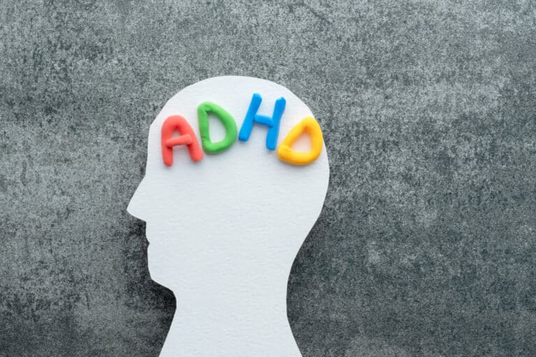 A white silhouette of a human head with colorful letters spelling "ADHD" placed on the brain area, set against a textured gray background.