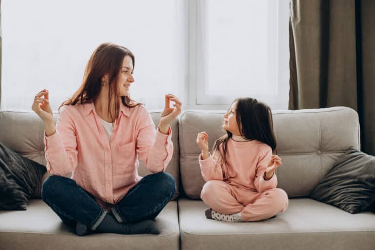 A mother and daughter sitting cross-legged on a sofa, smiling and practicing mindfulness together. Both are wearing pink pajamas in a cozy living room setting.