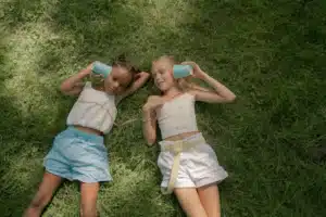 Two young girls lying on the grass using a string telephone with cups.