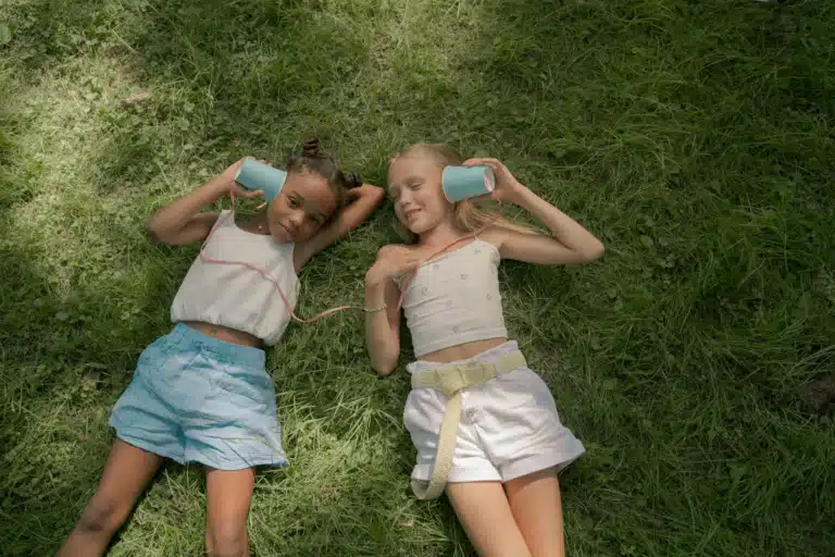 Two young girls lying on the grass using a string telephone with cups.