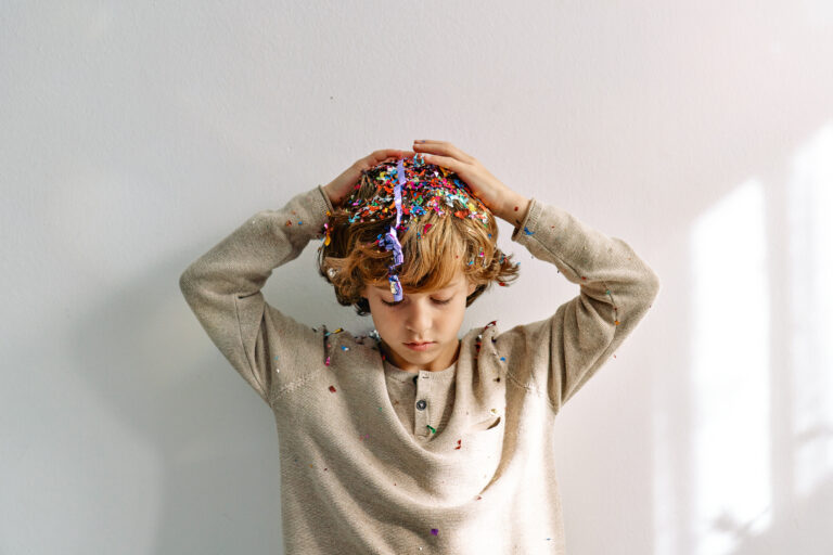 A young boy with confetti on his head and clothes, holding his hands on his head, standing against a plain white background.