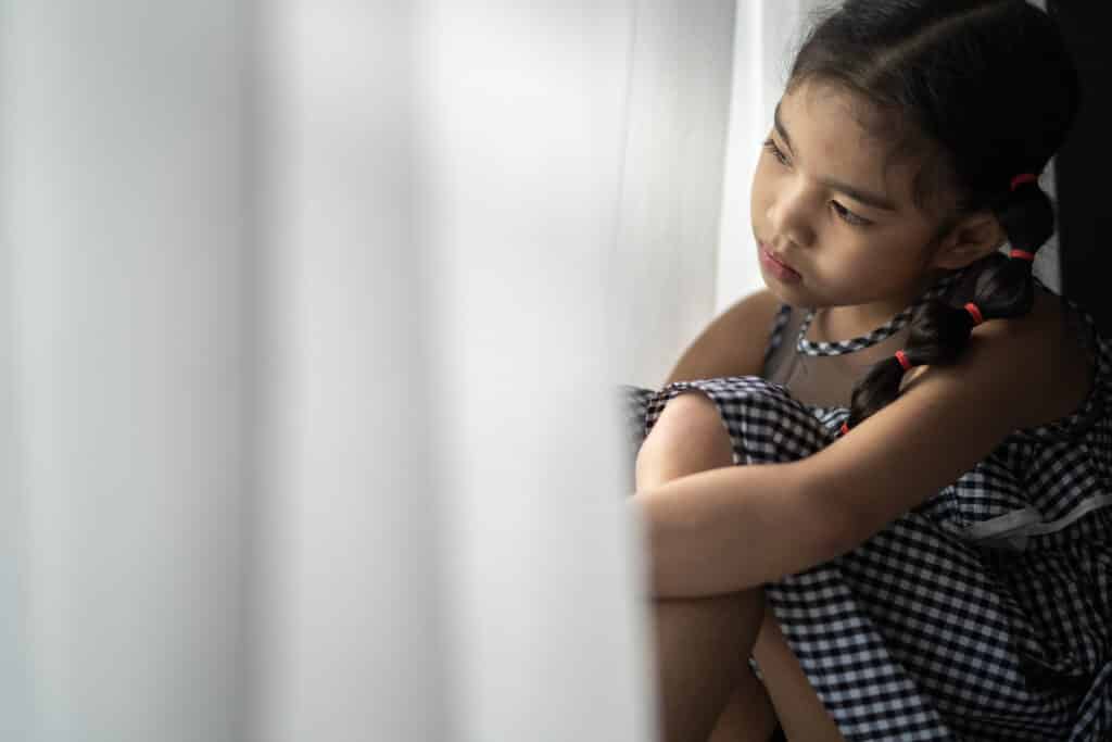 Sad young girl sitting by a window, looking outside thoughtfully.