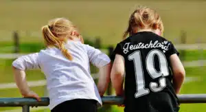 Two young girls leaning over a fence, one wearing a black shirt with "Deutschland 19" printed on the back.