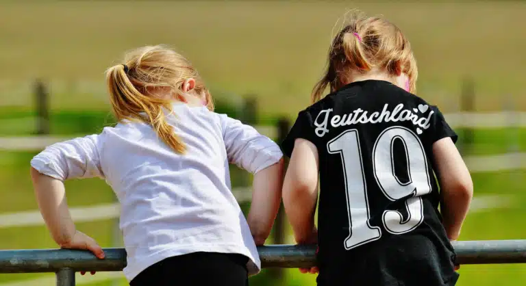 Two young girls leaning over a fence, one wearing a black shirt with "Deutschland 19" printed on the back.