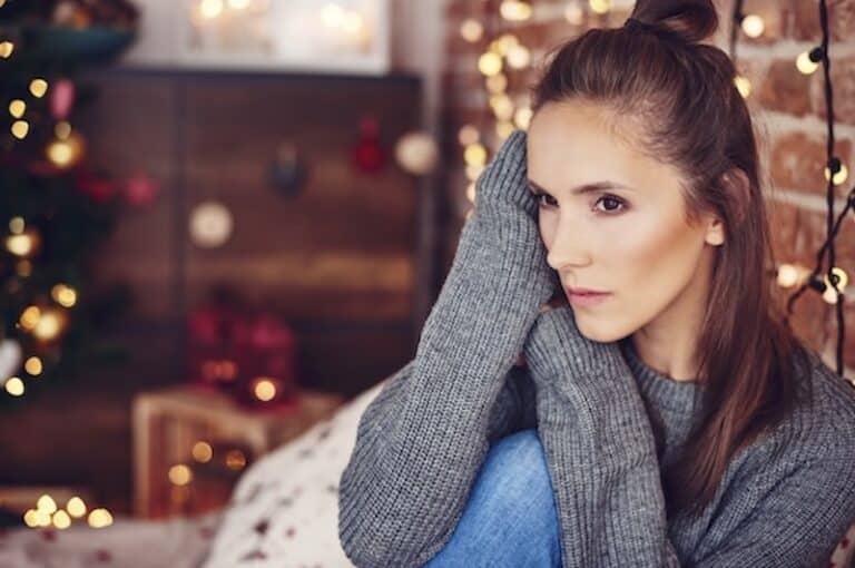 A thoughtful young woman sitting by a cozy fireplace with festive decorations in the background.