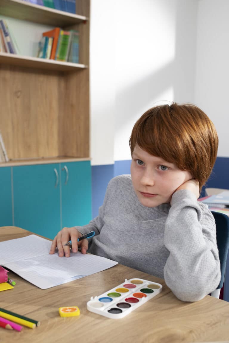 A young boy sitting at a desk in a classroom, appearing thoughtful or distracted, with school supplies like watercolor paints and paper in front of him.