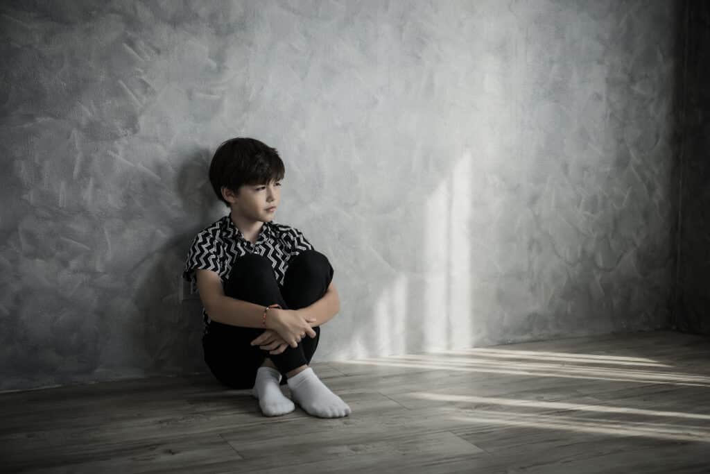 Sad boy sitting on the floor with arms wrapped around his knees, leaning against a gray wall.