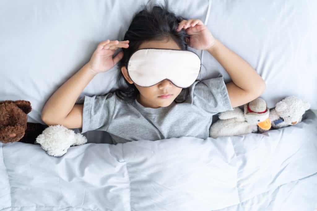 A young child wearing a sleep mask, lying in bed surrounded by stuffed animals.